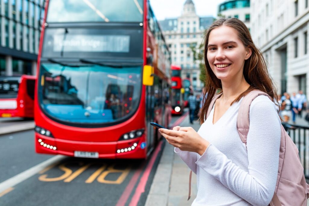 Smiling Woman With Smartphone At Bus Stop In London 1024x682